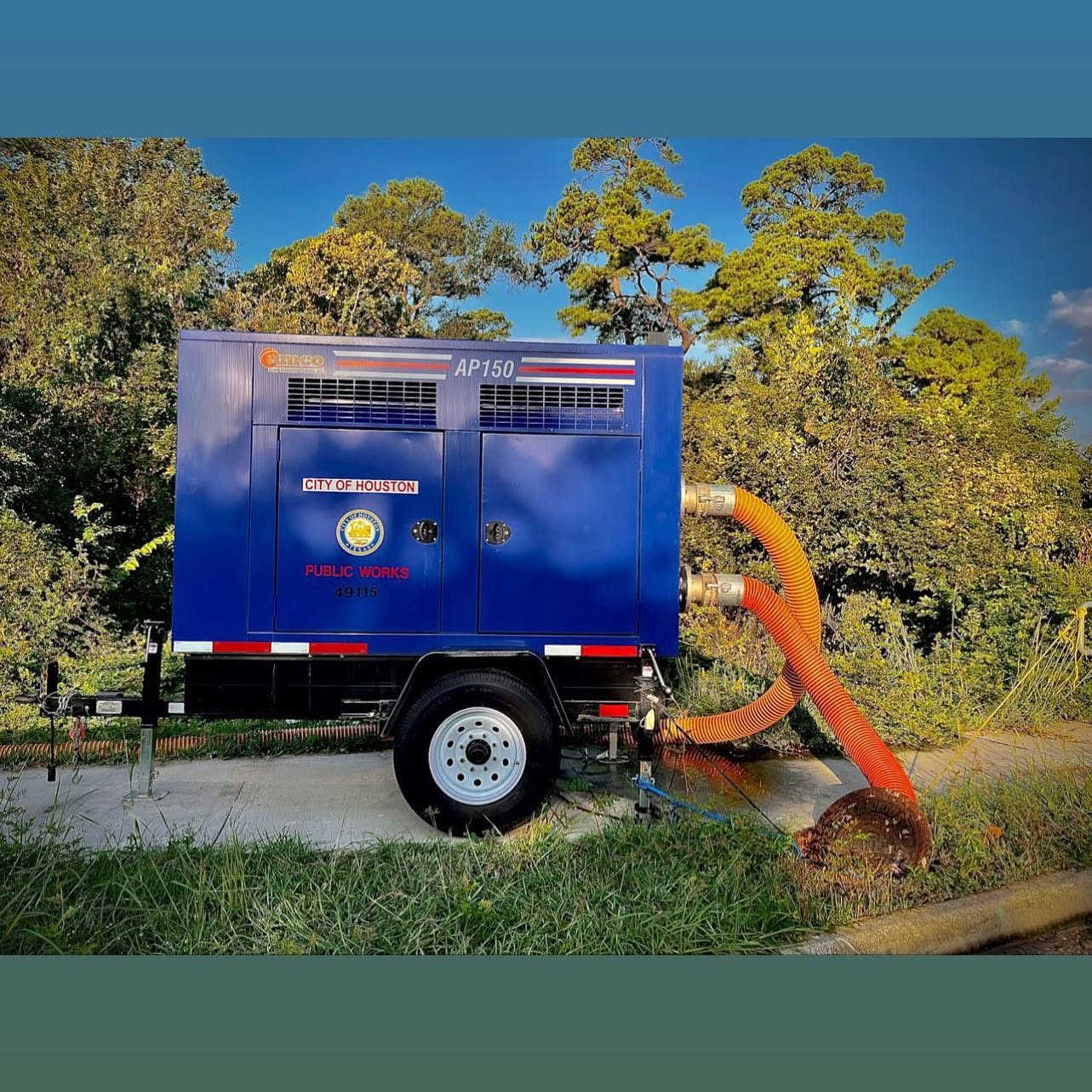 Blue AMCO AP150 sound-attenuated trailer-mounted pump marked City of Houston Public Works, with orange suction hose in a manhole at a construction site